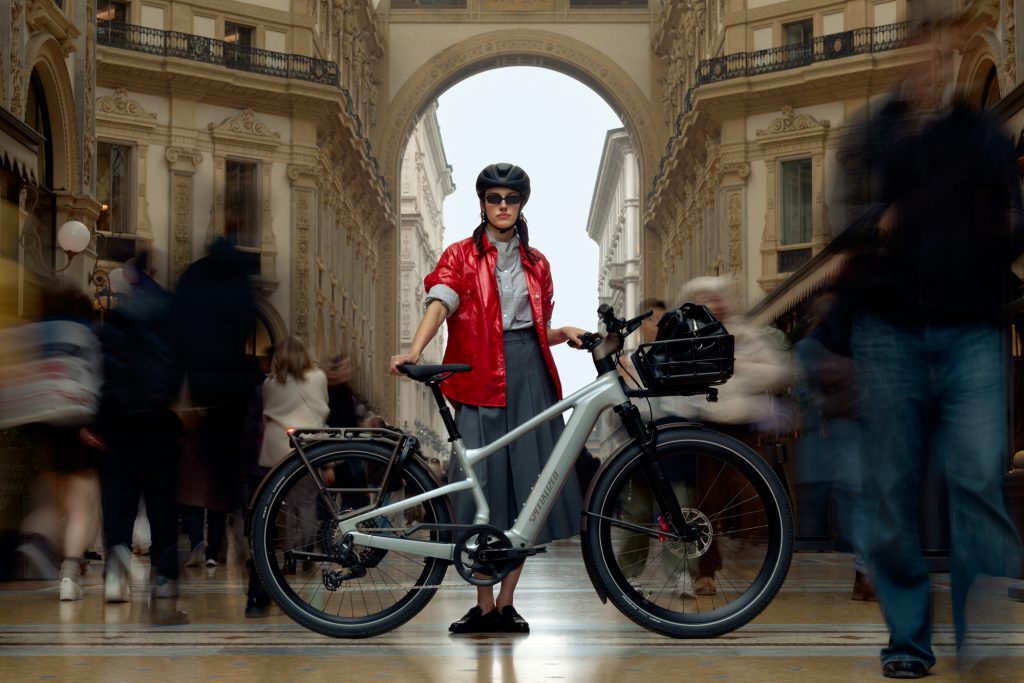 En person i röd jacka, hjälm och solglasögon står med en cykel i mitten av en stor, täckt shoppinggalleria. Oskarpa fotgängare rör sig runt dem, vilket framhäver kontrasten mellan stillhet och rörelse.