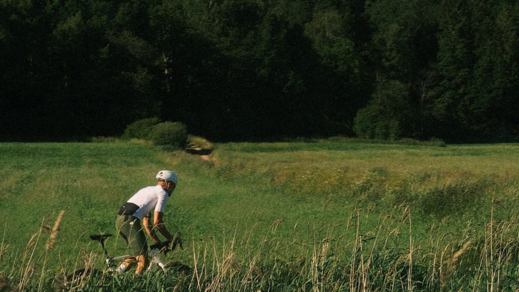 En person som bär hjälm och cykelkläder cyklar på en elcykel genom högt gräs på en grön, öppen äng med en tät skog i bakgrunden.