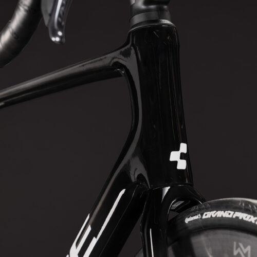 Närbild av den främre delen av en Racer Cube Attain C:62 SLT, Ultegra Di2 – Svart landsvägscykel med en vit logotyp, där styret, styrröret och en del av framhjulet framhävs med texten ”Grand Prix 5000” mot en mörk, enfärgad bakgrund.