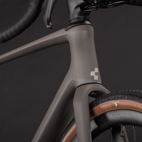 Närbild på en mattbrun Gravel Cube Nuroad C:62 Pro, en cykelram i 2x12s-konfiguration, där styrröret och framhjulet framhävs, med drop-styre och en minimalistisk vit logotyp som syns mot en svart bakgrund.