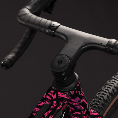 Närbild på en Gravel CUBE Nuroad C:62 SLX, 1x12s – Rosa cykelstyre och styrstam insvepta i svart tejp, monterade på en ram med ett iögonfallande geometriskt mönster i svart och rosa. Bakgrunden är mörk och mjukt suddig.