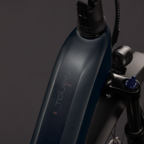 Närbild av det mörkfärgade batteriet som är monterat på ramen på en EL-cykel Cube Touring Hybrid ONE 600 Lågt insteg – Grå elcykel, med en synlig ”TOURING”-etikett. En del av framgaffeln, däcket och fjädringen syns också mot en mörk bakgrund.