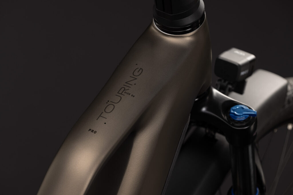 Närbild på den mattbronsfärgade ramen på EL-cykel Cube Touring Hybrid Pro 800, Step Trough – Brun, med texten ”TOURING PRO” nära styrlagret; en del av styret, framgaffeln och en monterad elektronisk enhet syns mot en mörk bakgrund.