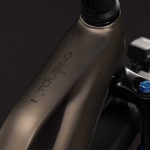 Närbild på den mattbronsfärgade ramen på EL-cykel Cube Touring Hybrid Pro 800, Step Trough – Brun, med texten ”TOURING PRO” nära styrlagret; en del av styret, framgaffeln och en monterad elektronisk enhet syns mot en mörk bakgrund.