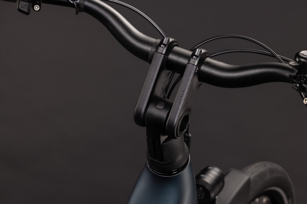 Närbild på styret och styrstammen på EL-cykel Cube Supreme RT Hybrid Comfort EX 600, Fotbroms – Blå/grön mot en enfärgad mörk bakgrund, där cykelramens främre del, bromskablarna och framhjulets ovansida syns.