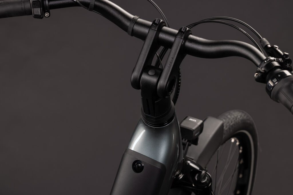 Närbild på styret, styrstammen och framhjulet på EL-cykel Cube Supreme RT Hybrid Deluxe EX 600, där den eleganta silvermetallic-ramen och de synliga bromskablarna framträder tydligt mot en mörk bakgrund.
