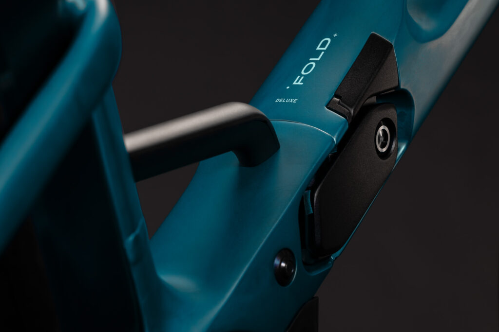 Närbild på ramen till en blå EL-cykel av modellen Cube Fold Hybrid Delu545, där den eleganta ”Fold Deluxe”-etiketten och den karakteristiska svarta låsmekanismen syns tydligt. Den mörka bakgrunden framhäver ramens släta yta och designelement, med subtila detaljer som antyder den innovativa Fold Hybrid-konstruktionen.