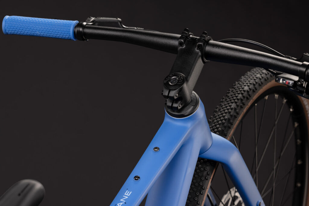 Närbild av Barncykel Cube Nulane Rookie ONE 28″ 1x9s – Blås mattblå styre, styrstam och framdäck mot en svart bakgrund, där en del av ramen, det strukturerade blåa styrehandtaget och däckets knubbiga mönster framhävs.