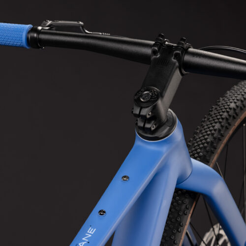Närbild av Barncykel Cube Nulane Rookie ONE 28″ 1x9s – Blås mattblå styre, styrstam och framdäck mot en svart bakgrund, där en del av ramen, det strukturerade blåa styrehandtaget och däckets knubbiga mönster framhävs.