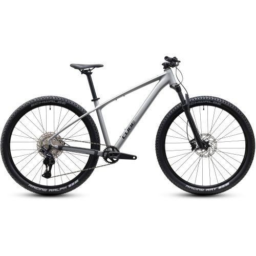 En silverfärgad mountainbike med rakt överrör, platt styre, framgaffel med fjädring och stora knubbiga däck. Cykeln avbildas från sidan mot en enfärgad vit bakgrund.