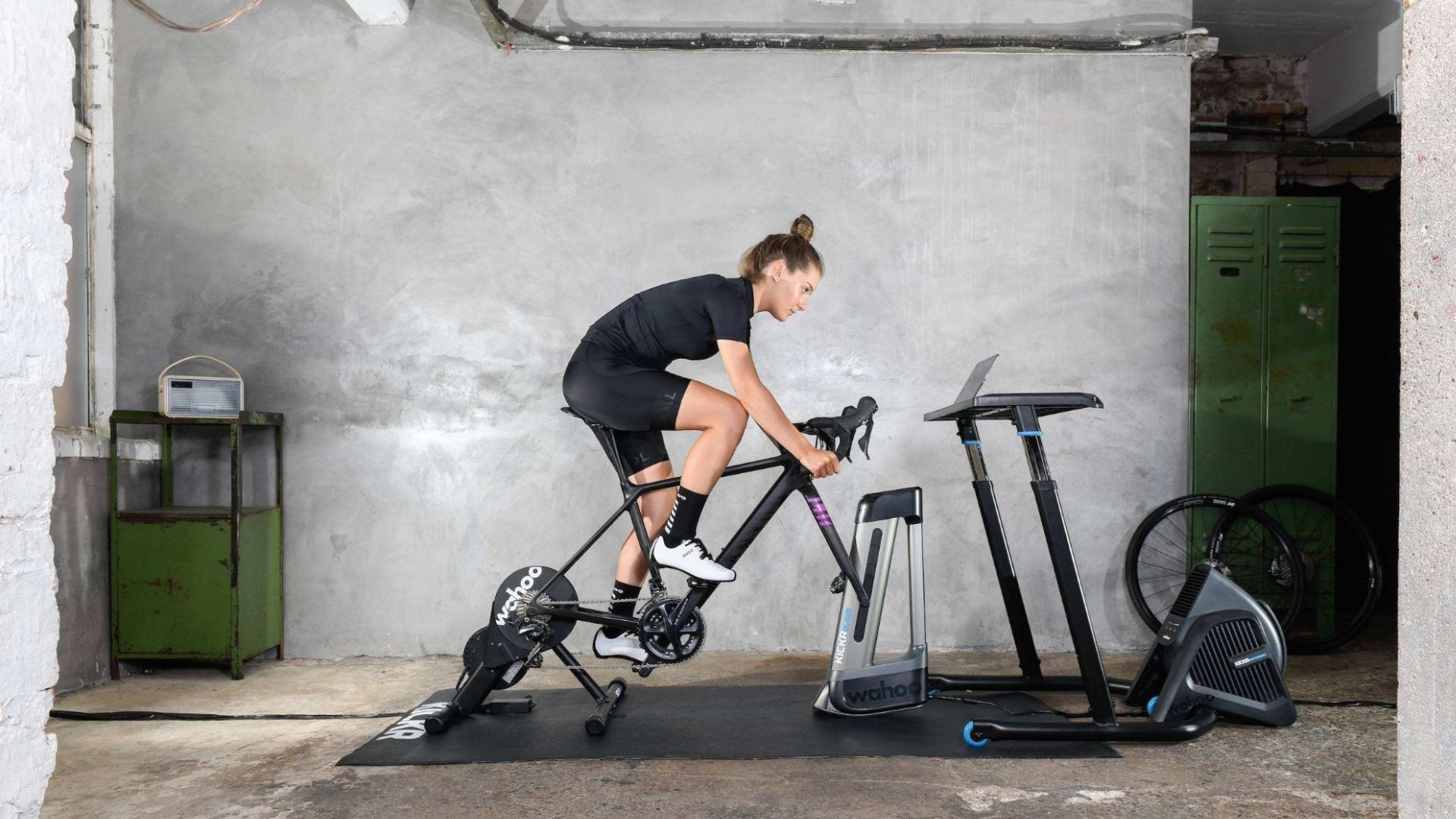 En person cyklar på en stationär cykel i ett betongrum, vänd mot en skärm som står på ett stativ. I närheten finns träningsredskap och ett grönt metallskåp. Personen, som är klädd i cykelkläder från den lokala cykelbutiken, verkar vara mitt uppe i ett träningspass.
