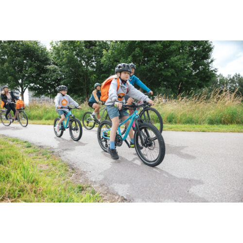 En grupp människor, bestående av både barn och vuxna, cyklar – till exempel på Crescent Njord 20″ Barncykel 3s i ljusblått eller beige – på en asfalterad cykelväg som kantas av gräs och träd. Alla bär hjälm och har ryggsäckar på sig. Vädret verkar vara milt och molnigt.