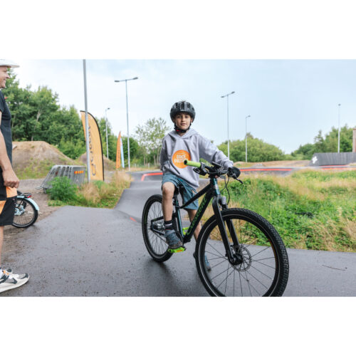 En pojke med hjälm och grå huvtröja cyklar på en Crescent Njord 24″ Barncykel 7s – Blå på en asfalterad stig i en utomhuspark. Till vänster syns en annan cykel och en del av en vuxen person. I bakgrunden finns gräsytor och gatlyktor.